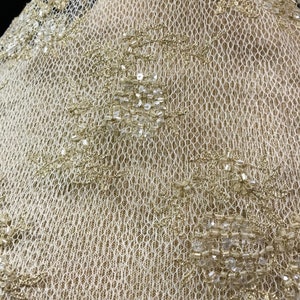 K&ouml;nnte beinhalten: Nahaufnahme eines cremefarbenen Spitzenstoffs mit aufwendigen Blumenmustern. Die Spitze ist mit goldfarbenen Perlen und Pailletten verziert, was einen schimmernden Effekt erzeugt. Die zarte netzartige Textur des Stoffes ist sichtbar.