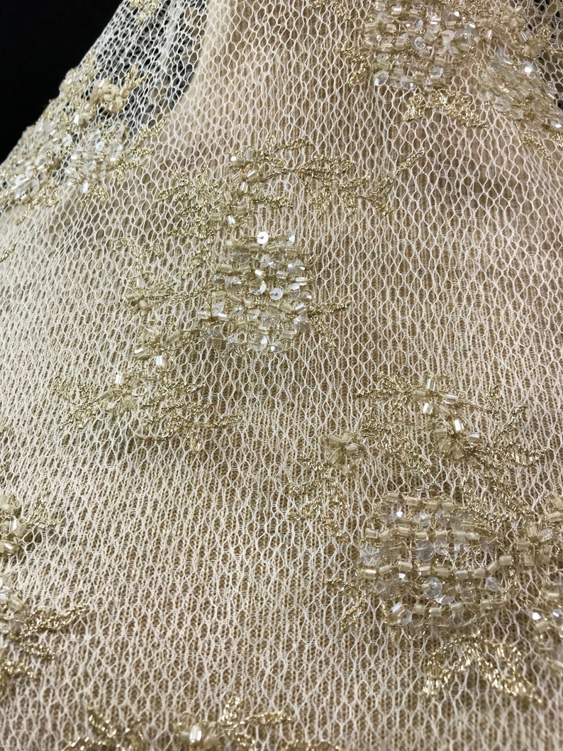 K&ouml;nnte beinhalten: Nahaufnahme eines cremefarbenen Spitzenstoffs mit aufwendigen Blumenmustern. Die Spitze ist mit goldfarbenen Perlen und Pailletten verziert, was einen schimmernden Effekt erzeugt. Die zarte netzartige Textur des Stoffes ist sichtbar.
