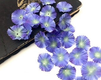 May include: A collection of small, artificial flowers with a blue-purple hue and a light green center. The flowers are scattered on a white surface and a dark, textured book. These decorative flowers are ideal for craft projects.