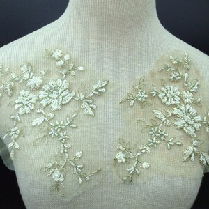 以下が含まれることがあります： 白と金の刺繍が施された2つの花のレースアップリケ。アップリケは、小さなビーズのアクセントが付いた、詳細な花と葉のデザインが特徴です。レースは、衣類や工芸品に最適な、透明なベージュのメッシュの裏地に付いています。