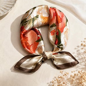May include: A square silk scarf featuring a vibrant floral pattern with large orange, red, and white blossoms set against a cream background. The scarf is tied in a knot and finished with a dark brown border that frames the colorful botanical design.