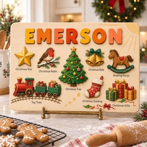 Può includere: Un puzzle rettangolare in legno con il nome EMERSON in grandi lettere colorate nella parte superiore. Include forme in legno dipinte e in rilievo di una stella, pettirosso, albero, campane, cavallo a dondolo, treno, calza e regali di Natale con etichette.