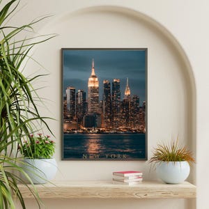 May include: A rectangular wall art print featuring a night cityscape of New York City with illuminated skyscrapers reflecting on the water. The text NEW YORK is printed in gold capital letters at the bottom centre of the dark blue and orange image.
