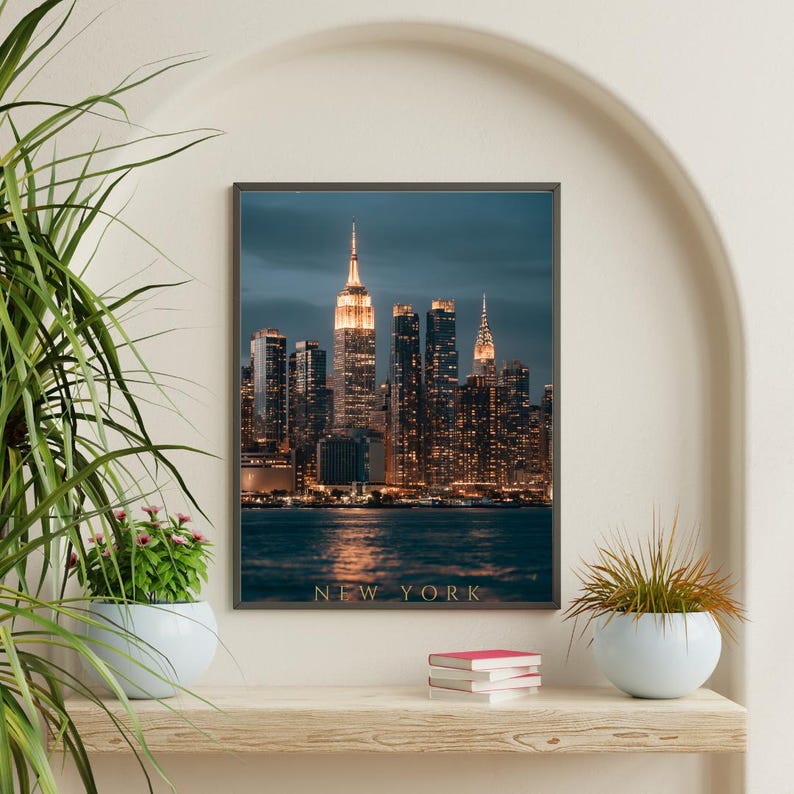May include: A rectangular wall art print featuring a night cityscape of New York City with illuminated skyscrapers reflecting on the water. The text NEW YORK is printed in gold capital letters at the bottom centre of the dark blue and orange image.