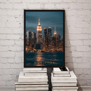 May include: A framed vertical wall art print featuring a night cityscape of New York City. The image displays illuminated skyscrapers, including the Empire State Building, reflecting on dark water. The text NEW YORK is printed in gold at the bottom.