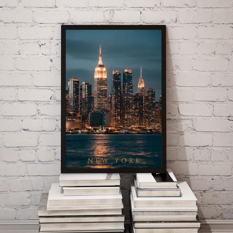 May include: A framed vertical wall art print featuring a night cityscape of New York City. The image displays illuminated skyscrapers, including the Empire State Building, reflecting on dark water. The text NEW YORK is printed in gold at the bottom.