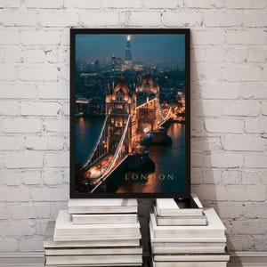 May include: A framed vertical wall art print featuring a night view of Tower Bridge in London, illuminated with warm golden lights against a dark blue twilight sky. The Shard skyscraper is visible in the distance. The word LONDON is printed in gold.