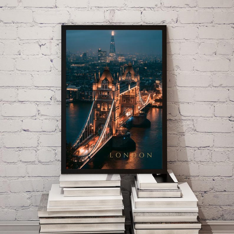 May include: A framed vertical wall art print featuring a night view of Tower Bridge in London, illuminated with warm golden lights against a dark blue twilight sky. The Shard skyscraper is visible in the distance. The word LONDON is printed in gold.