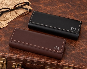 Puede incluir: Dos estuches rectangulares para gafas, uno negro y otro marrón oscuro, con costuras claras visibles en los bordes. El estuche negro está grabado con las iniciales DS y el estuche marrón oscuro con las iniciales JM.