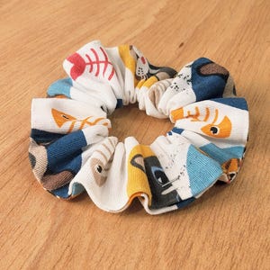 Puede incluir: Un coletero de tela con un patrón repetitivo de espinas de pescado y caras de gato coloridas sobre un fondo blanco. El coletero tiene una textura fruncida y plisada con detalles en azul, naranja, rojo y negro en todo el diseño estampado.