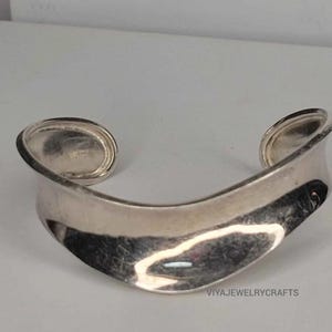 Può includere: Un bracciale a fascia in argento lucido con un design ampio, curvo e ondulato. La superficie metallica è liscia e riflettente, caratterizzata da una struttura a fascia aperta continua. Il testo VIYAJEWELRYCRAFTS è stampato nell'angolo in basso a destra.