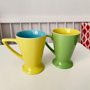 Könnte beinhalten: Zwei Keramikbecher mit ausgestellten Rändern und Sockelfüßen. Der linke Becher ist leuchtend gelb mit hellblauem Innenraum und einem eckigen Henkel. Der rechte Becher ist lindgrün mit gelbem Innenraum und einem passenden grünen Henkel.