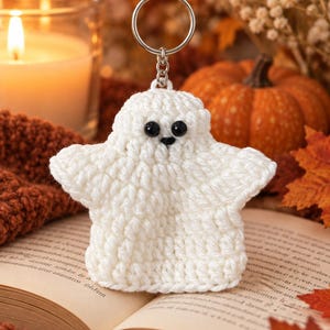 May include: A white crocheted ghost keychain featuring two small black plastic eyes and a tiny black stitched nose. The ghost is crafted from yarn with a textured surface and is attached to a circular silver metal split ring by a short chain.