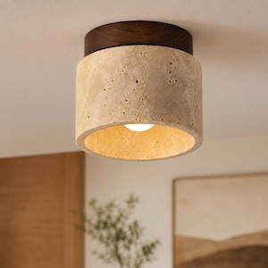 Pode incluir: Uma luminária de teto embutida com uma cúpula cilíndrica feita de pedra travertino bege porosa. A parte superior da luminária consiste em uma base circular de madeira marrom escura que se fixa diretamente à superfície do teto.