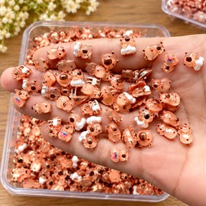 May include: A collection of small, rose gold-toned metal charms shaped like stylized bears. Each charm features a smooth, metallic finish with white enamel accents on the head and body, and a single dark, circular eye detail.