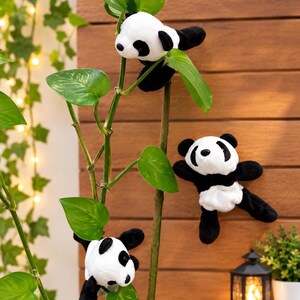 May include: Three plush panda toys with black and white fur, each featuring round black ears, black eye patches, and black limbs. The stuffed animals are positioned on the stems and leaves of a green plant, showcasing their soft, fuzzy texture.