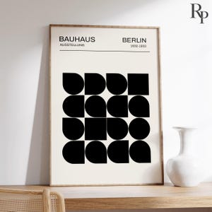 以下が含まれることがあります： 上部に「BAUHAUS AUSSTELLUNG BERLIN 1932-1933」の文字が記された額入りポスター。テキストの下には、クリーム色の背景に円、正方形、扇形などの黒い幾何学模様が配置された16個の正方形のグリッドが描かれています。