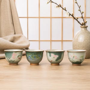 Puede incluir: Cuatro pequeñas tazas de té de cerámica dispuestas en fila, cada una con un diseño pintado a mano de flores de loto y hojas verdes sobre un esmalte craquelado. Las tazas tienen una paleta de colores verde claro y crema.