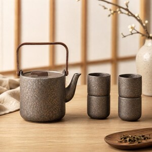 Puede incluir: Una tetera de cerámica gris oscuro con acabado texturizado, tapa plana y asa basculante de madera marrón, acompañada de cuatro tazas de té cilíndricas a juego apiladas en dos pares. El conjunto presenta una textura moteada en toda su superficie.