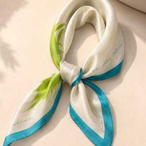 May include: A square silk scarf featuring a cream-colored base with a vibrant green palm leaf pattern on one side and a solid teal border. The fabric has a smooth, lustrous finish and includes the cursive text fashion printed in a dark script.