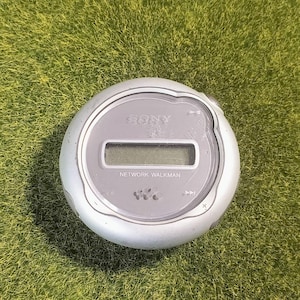 May include: A circular silver Sony Network Walkman digital music player featuring a central rectangular LCD screen, playback control icons, and the Sony logo printed on the top surface of the device casing with a smooth, metallic finish.