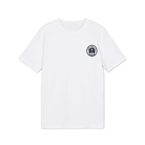 Puede incluir: Una camiseta blanca de manga corta con un pequeño gráfico circular en el pecho izquierdo. El gráfico muestra una gorra de béisbol azul marino con el texto NEW YORK CITY y BASEBALL en un borde circular con dos bates de béisbol cruzados.