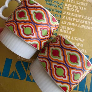 May include: Two ceramic mugs featuring a repeating geometric pattern in orange, purple, lime green, and white. The mugs have a white ribbed base and a heart-shaped handle. The upper portion of each mug displays a vibrant, interlocking tile-like design.