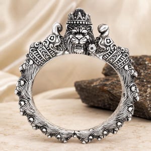 Puede incluir: Un brazalete circular de tono plateado que presenta un motivo prominente de cabeza de león en la parte superior, adornado con una pequeña corona. La banda está decorada con intrincados patrones en relieve texturizados alrededor de toda la circunferencia.