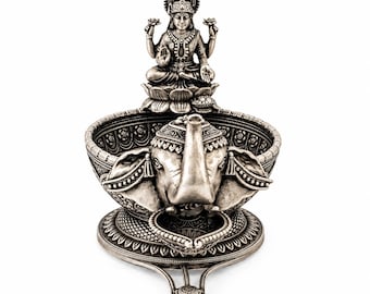 May include: A silver-toned metal oil lamp featuring a central elephant head sculpture and a seated deity figure positioned above the bowl. The bowl is decorated with intricate floral patterns and rests on a circular base with a small protruding spout.