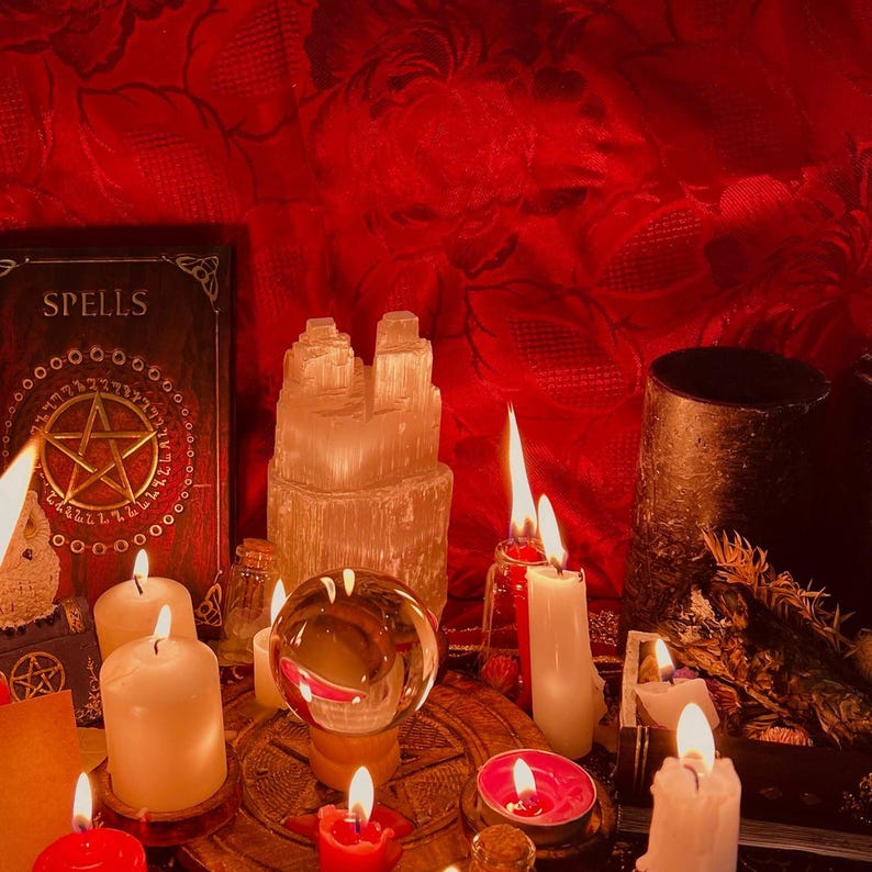 May include: A collection of ritual items including a book titled SPELLS with a gold pentagram, a clear crystal sphere on a wooden base, a tall selenite tower, and multiple lit white and red candles. The scene is set against a deep red fabric.