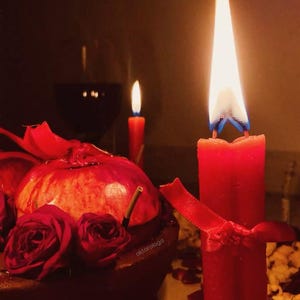 May include: A tall red candle with a bright flame, tied with a red ribbon, stands next to a dark bowl containing a pomegranate and deep red roses. A second, smaller red candle burns in the background, alongside a glass of dark liquid.