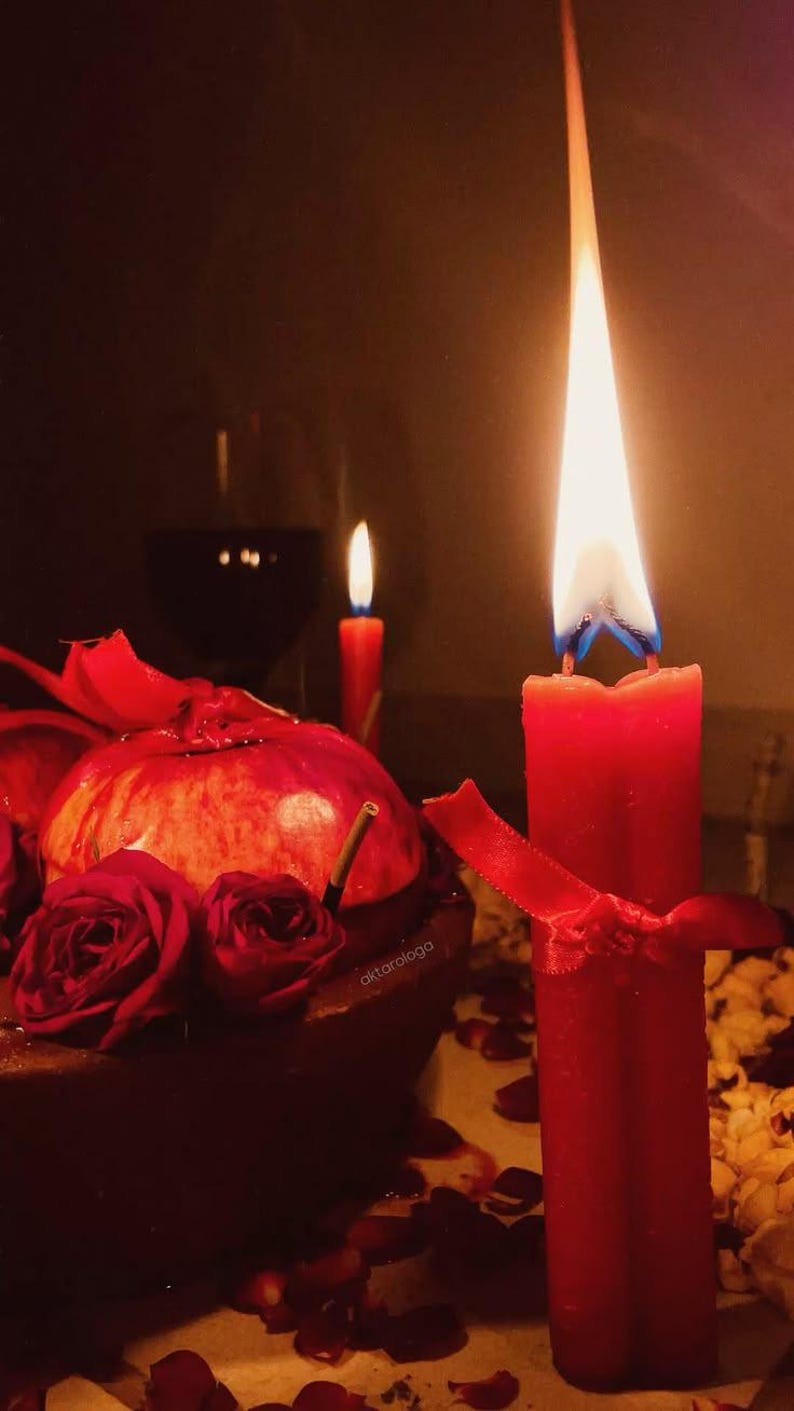 May include: A tall red candle with a bright flame, tied with a red ribbon, stands next to a dark bowl containing a pomegranate and deep red roses. A second, smaller red candle burns in the background, alongside a glass of dark liquid.