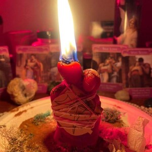 May include: A red candle with a bright flame, wrapped in a piece of paper secured with thin white string. The candle is positioned on a surface covered with scattered dried herbs and plant matter, illuminated by a warm, reddish light.