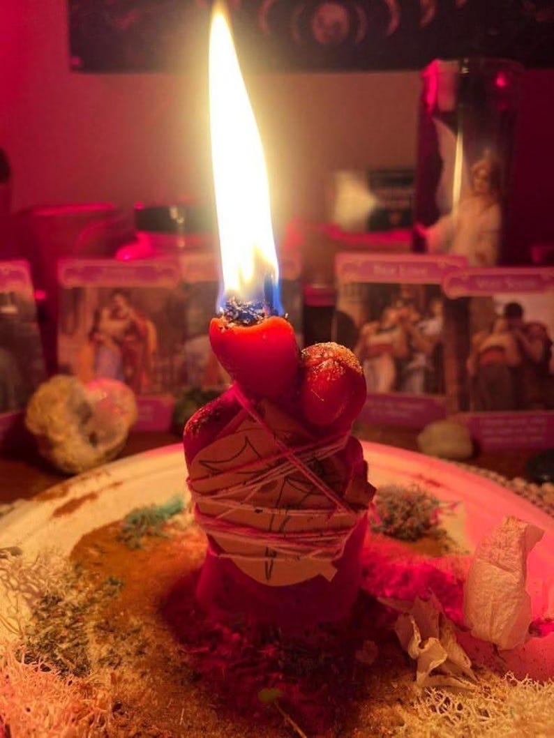 May include: A red candle with a bright flame, wrapped in a piece of paper secured with thin white string. The candle is positioned on a surface covered with scattered dried herbs and plant matter, illuminated by a warm, reddish light.