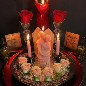May include: A centerpiece featuring a pink candle shaped like two figures embracing, surrounded by light pink roses, two lit taper candles in black holders, and two red roses in glass vases. Two tarot cards and two glass dropper bottles are present.