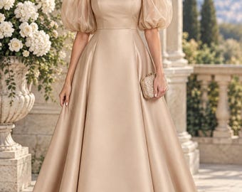 May include: A floor-length champagne-colored gown featuring a square neckline, voluminous sheer puff sleeves, and a full A-line skirt. The dress is crafted from a smooth, satin-like fabric and is paired with a small, rectangular, embellished clutch.