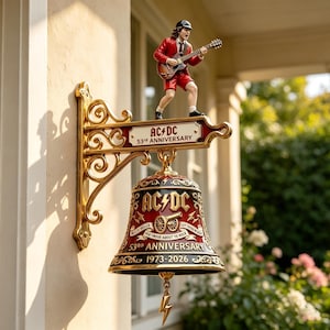 Könnte beinhalten: Eine dekorative Wandglocke mit einer goldfarbenen, verzierten Halterung und einer dunkelroten Glocke mit goldenen Akzenten. Die Glocke zeigt den Text AC/DC, 53rd ANNIVERSARY und 1973-2026. Eine Gitarristenfigur im roten Anzug steht auf der Halterung.