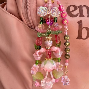 May include: A decorative keychain featuring a central fairy-like figure crafted from pink and green beads, accented with iridescent rose-shaped beads, a heart-shaped bead, and a dangling strand of letter beads spelling out the word DEW DROP.