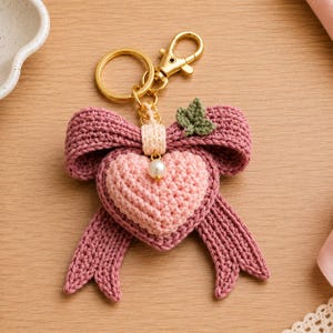 May include: A crochet keychain featuring a dusty rose heart centered on a larger mauve bow. The design includes a small cream-colored center band with a white pearl bead, a tiny green leaf accent, and a gold-toned metal ring with a lobster clasp.