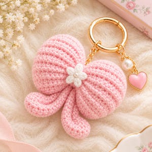 Puede incluir: Un llavero de lazo tejido a ganchillo en color rosa claro con una pequeña flor blanca tejida en el centro. El llavero incluye una anilla de metal dorado, una cadena corta, una cuenta de perla blanca y un pequeño dije de corazón rosa.