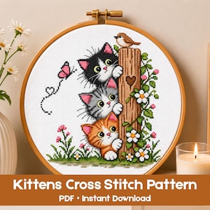 May include: A cross stitch pattern in a wooden hoop featuring three stacked kittens—black and white, gray tabby, and orange tabby—peeking from behind a wooden post with a small bird on top, a pink butterfly, and white flowers on green stems.