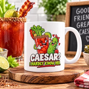 May include: A white ceramic mug featuring a cartoon illustration of a Caesar cocktail and a pickle character. The text reads "CAESAR? I HARDLY KNOW HER." The design includes red, green, and brown colours, depicting a drink with celery and bacon.