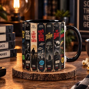 May include: A black ceramic mug featuring a wrap-around graphic of vintage horror movie VHS tape spines. The design includes titles like Night of the Living Dead, The Last Man on Earth, Plan 9 from Outer Space, The Terror, The Bat, and Little Shop of Horrors.
