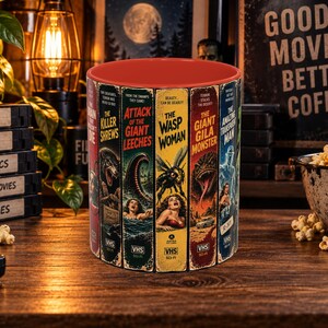 May include: A ceramic mug with a red interior and a wrap-around design featuring vintage VHS tape covers for sci-fi horror films including The Killer Shrews, Attack of the Giant Leeches, The Wasp Woman, The Giant Gila Monster, and The Amazing Transparent Man.