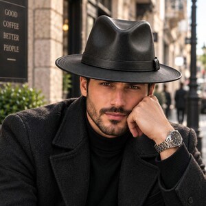 Pode incluir: Um chapéu fedora preto com uma copa estruturada e aba larga, apresentando uma faixa preta a condizer à volta da base. O chapéu é feito de um material liso e é usado com uma camisola de gola alta preta e um casaco preto com um relógio.