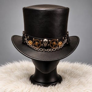May include: A black top hat featuring a wide band adorned with a central silver-toned skull, two bronze-coloured gear accents, and a silver metal chain draped across the front. The hat has a smooth, matte finish and a structured, curved brim.