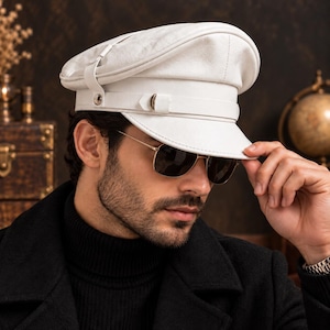 Pode incluir: Um boné militar de couro branco com uma coroa estruturada, uma pala plana e uma tira horizontal decorativa na frente, fixada com ferragens de metal prateado. O boné tem um acabamento liso e uma silhueta clássica de pala.