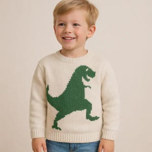 May include: A cream-colored long-sleeve knit sweater featuring a large, dark green graphic of a Tyrannosaurus Rex dinosaur centered on the front. The sweater has a ribbed crew neckline, ribbed cuffs, and a ribbed hem, providing a classic fit.