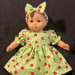 May include: A green and red ladybug print dress with white lace trim and matching headband for a doll. The doll is wearing white shoes.