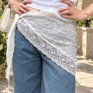 May include: A white sheer fabric wrap featuring intricate floral embroidery and a wide scalloped lace border. The wrap is tied around the waist over blue denim jeans, paired with a white top and a cream-colored handbag with gold hardware.